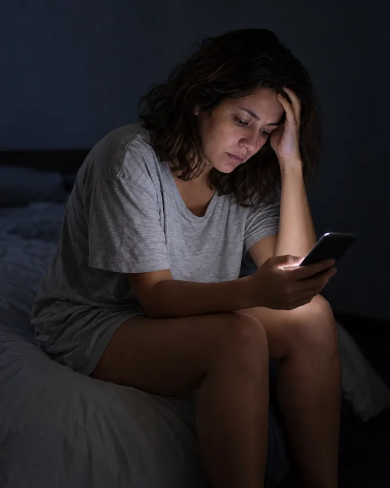Mulher sentada na cama de madrugada, olhando o celular com cara de cansaço.